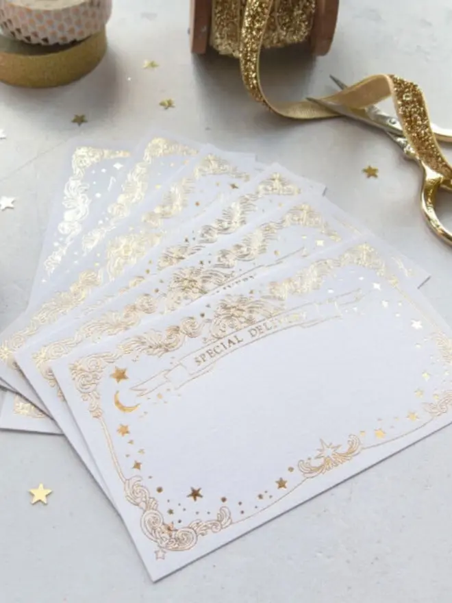 A fan of blank white "Special Delivery" Christmas notelets with shimmering gold foil borders of scrolls, stars and a crescent moon, beside gold glitter ribbon, washi tape, gold scissors and scattered star confetti.