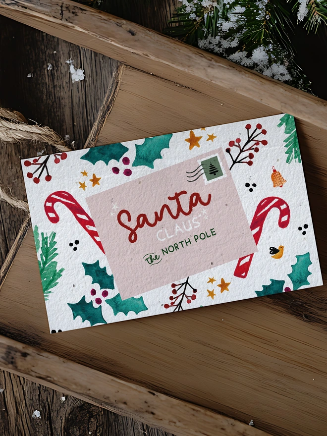 A Christmas card with an illustrated postcard addressed to Santa Claus with candy canes and holy leaves. The card is on a wooden sleigh with greenery