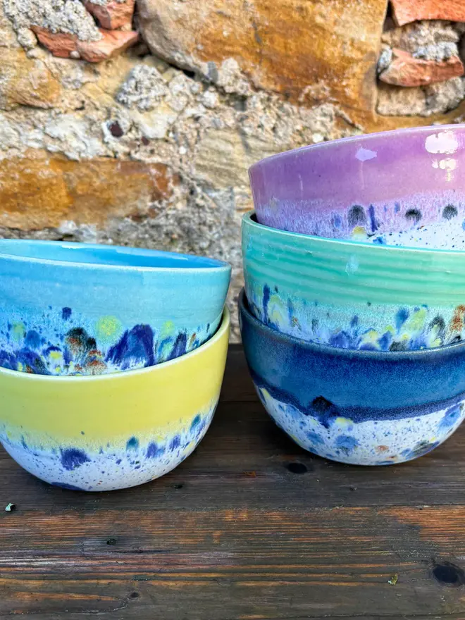 Shoreline Small Bowl, a group of colourful bowls stacked on top of each other on a wooden surface in front of a brick wall. 