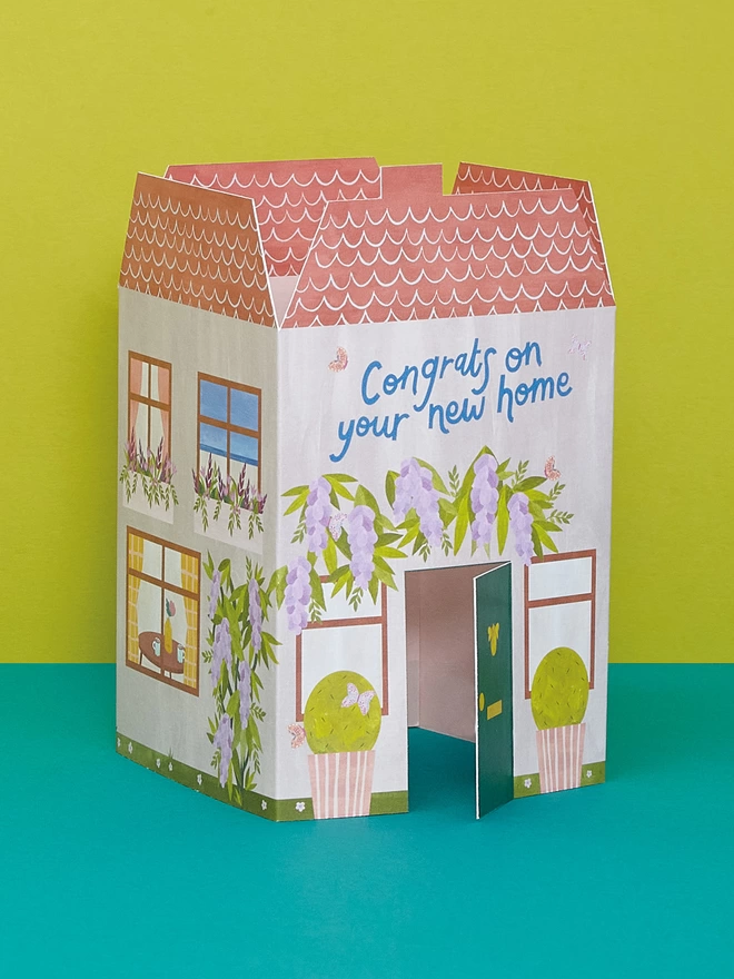 3D fold out house with die cut door that opens out. The house has painted climbing wisteria, colourful windows and plants, and blue hand lettering with a 'Congrats on your new home' message