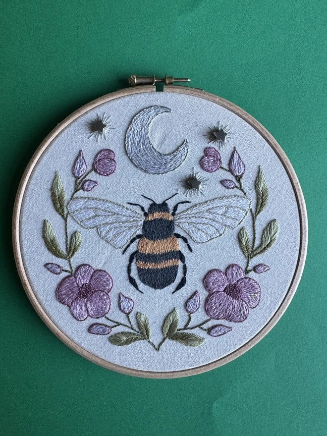 An embroidery hoop on a green background containing a stitched bee pink flowers green leaves a silver crescent moon and star shaped beads