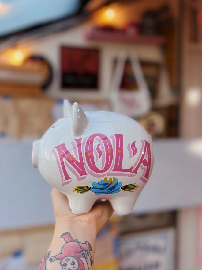 White piggy bank painted with 'NOLA' in serif lettering, in a hot pink with pale pink shade, and a blue canal rose and green leaves.