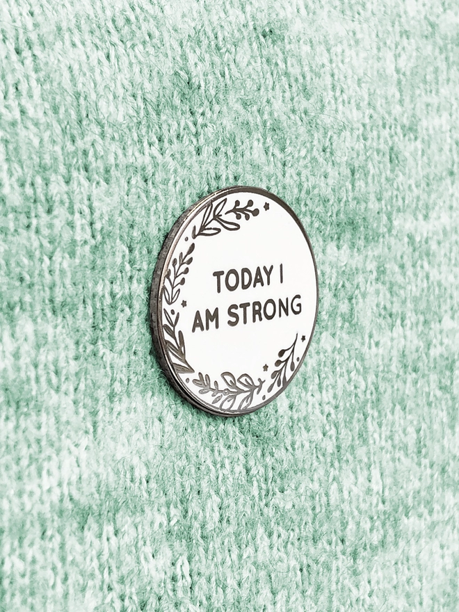 A round white enamel pin with a floral design and the words "Today I Am Strong" is pinned to green fabric.