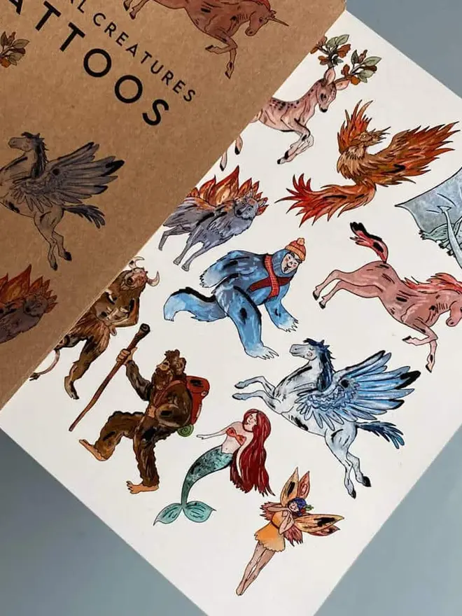 Dinosaur & Fantasy Temporary Tattoos, A brown kraft paper booklet titled "MYTHICAL CREATURES TATTOOS" sits next to a sheet of colorful mythical creature temporary tattoos.