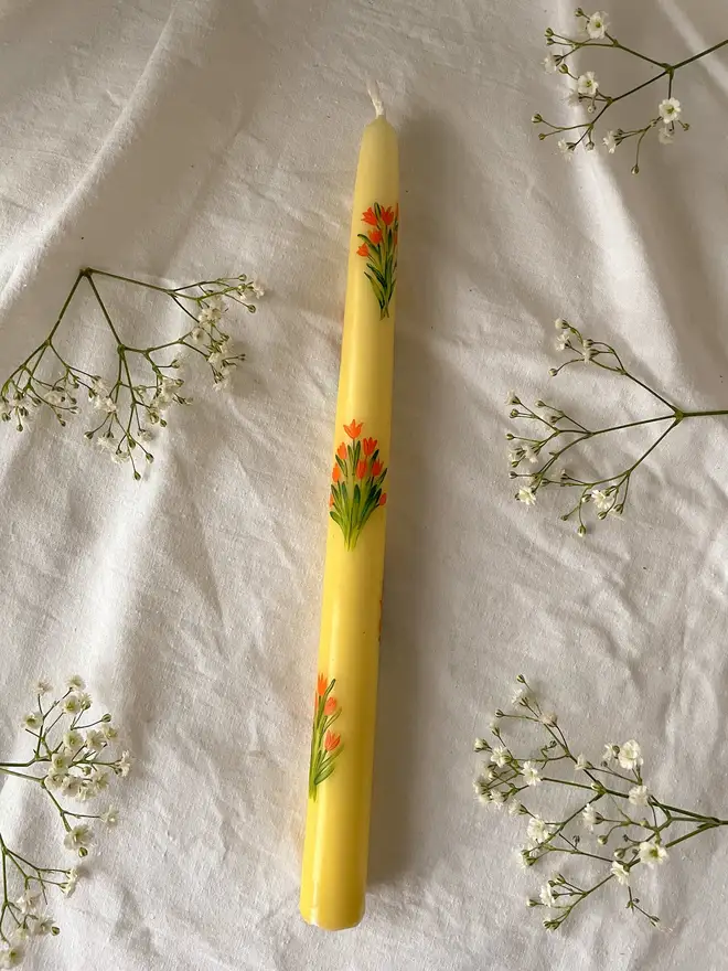 Mon Cherry & Tulip Fever Candle, a yellow candle lying on a table cloth accompanied by flowers. It's floral design wraps around the stalk. 