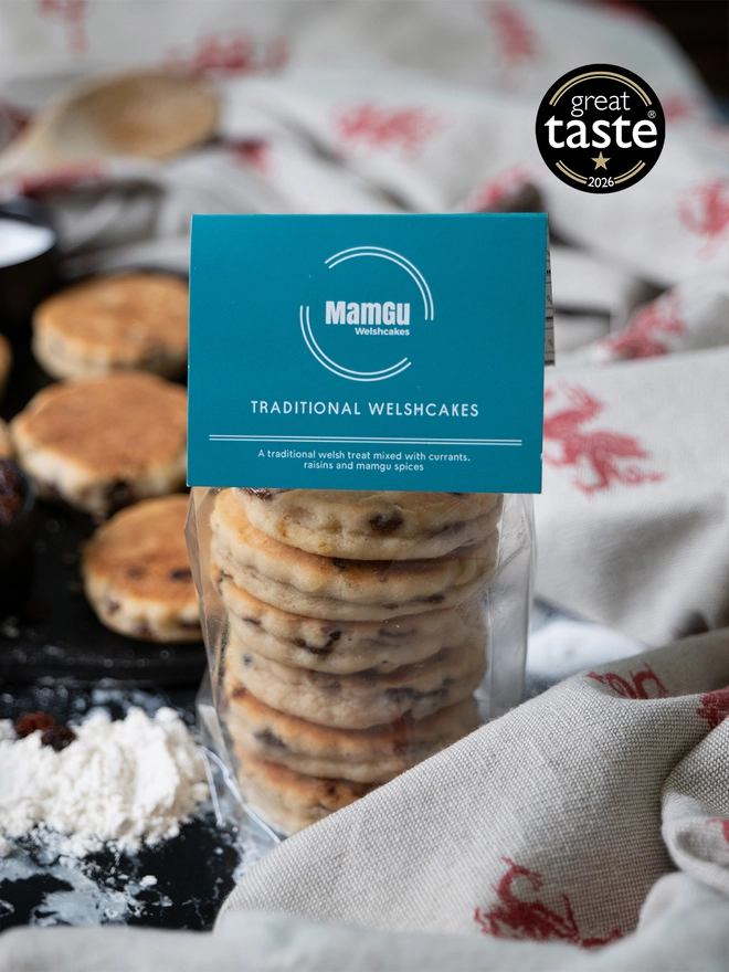 A pack of 6 Traditional Welsh Cakes with a teal header card, sitting on a kitchen work top with loose welsh cakes and ingredients