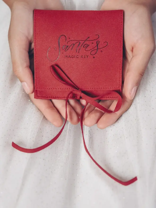 Cupped hands hold a red suede pouch printed with the "Santa's Magic Key" logo. A matching red ribbon is tied in a bow at the front.
