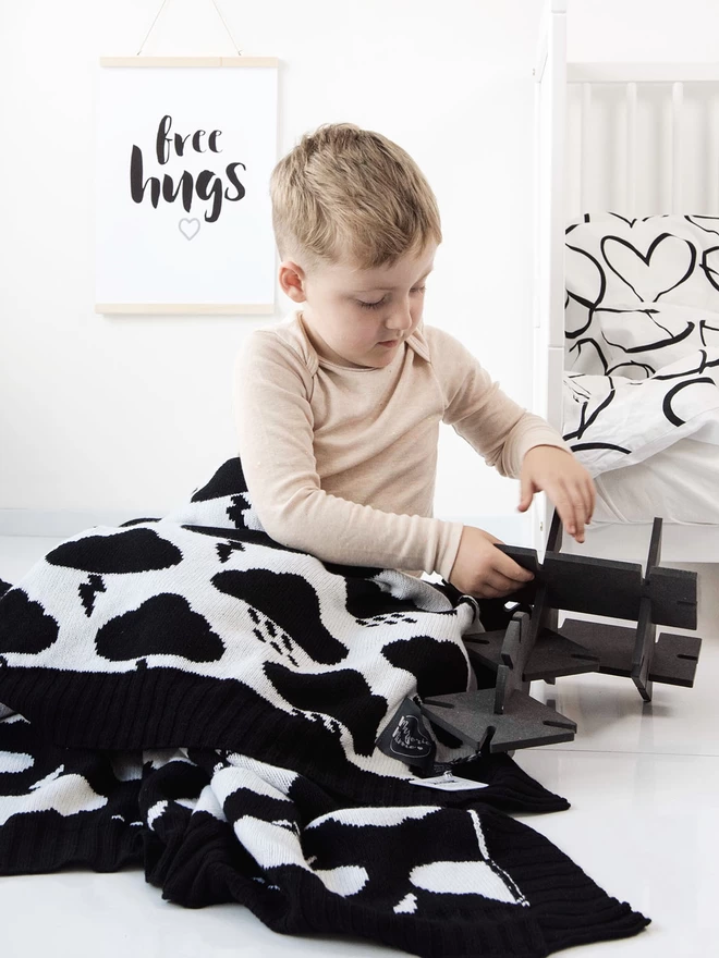 A young boy sits on the floor playing with a wooden puzzle.  He is wearing his pyjamas and a bed with a black and white heart duvet on is visible in the background. On his knees is a black and white storm cloud blanket and a monochrome print behind his head read 'free hugs'.