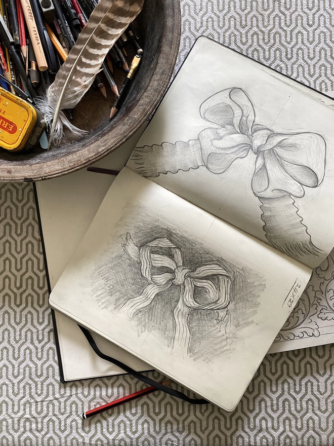 Sketchbook with sketches of ribbons and bows and a wooden bowl of drawing equipment