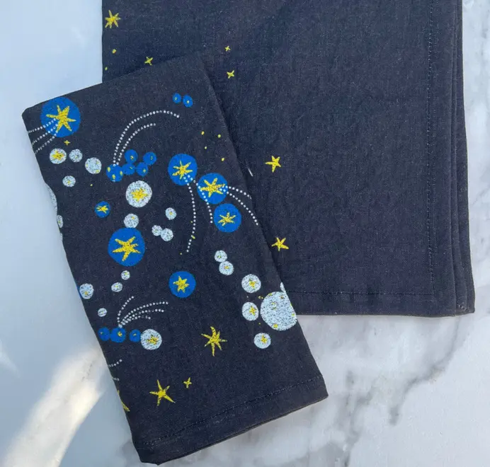 Solaris Napkins In White Or Blue. Blue cloth napkin with blue and yellow starburst and orbital patterns. 