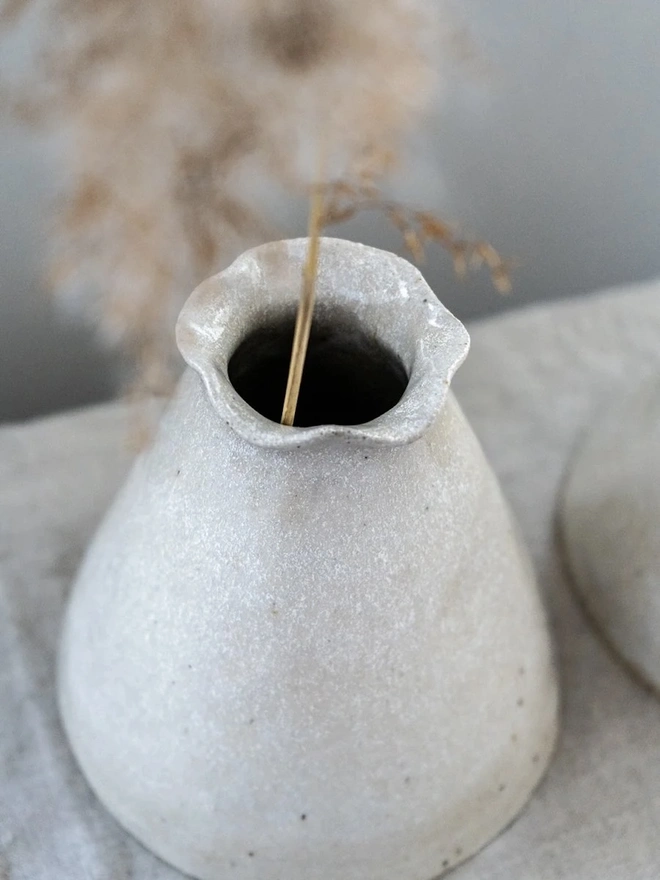 Maia Bud Vase In Sea Salt Glaze, with plant in it, placed on a table cloth against a soft background. 