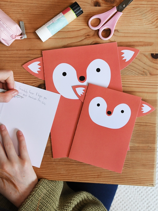 Several orange fox greetings cards lay on a wooden table. A child is writing a message in one of the cards.