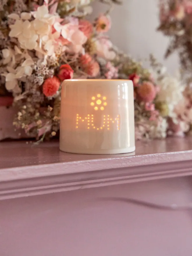 Porcelain tea light holder with 'MUM' and a flower detail - a tea light lit inside, on a pink mantlepiece surrounded by flowers
