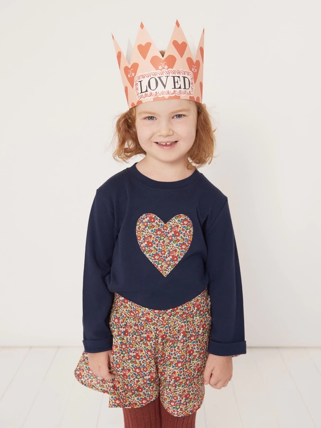 Smiling girl wearing a navy t-shirt with a floral heart sewn on the front and a crown that says love
