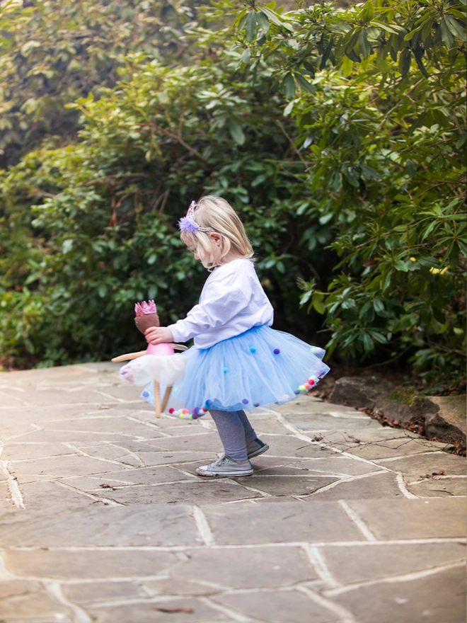 Little girl in a park twirling while holding a doll and wearing a baby blue pom pom tutu 