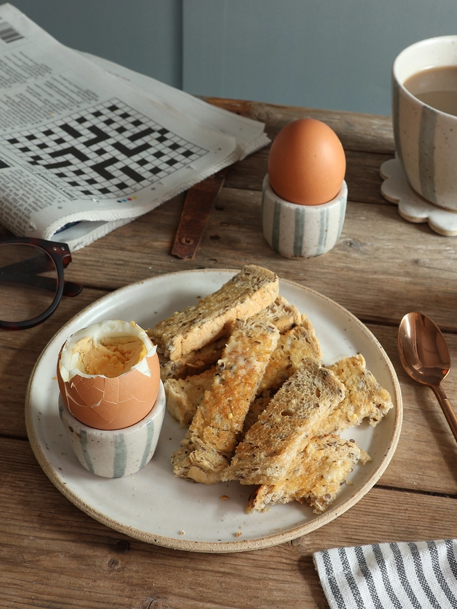 A breakfast scene featuring two handmade ceramic egg cups with a blue and white stripe design holding boiled eggs. One of the egg cups sits on a white handmade ceramic plate filled with buttered toast. A newspaper, cloth napkin, brass spoon and blue and white striped mug also sit on the table. 