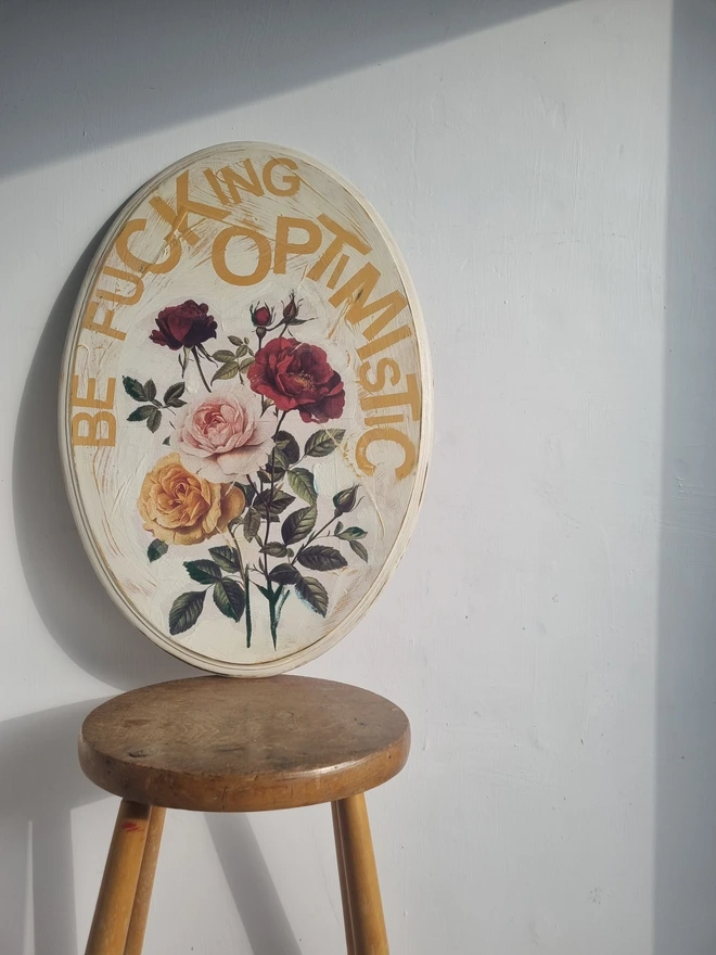 An art panel made from an upcycled table top. The image is vintage roses on a cream background with text that reads 'be fucking optimistic' in mustard yellow..