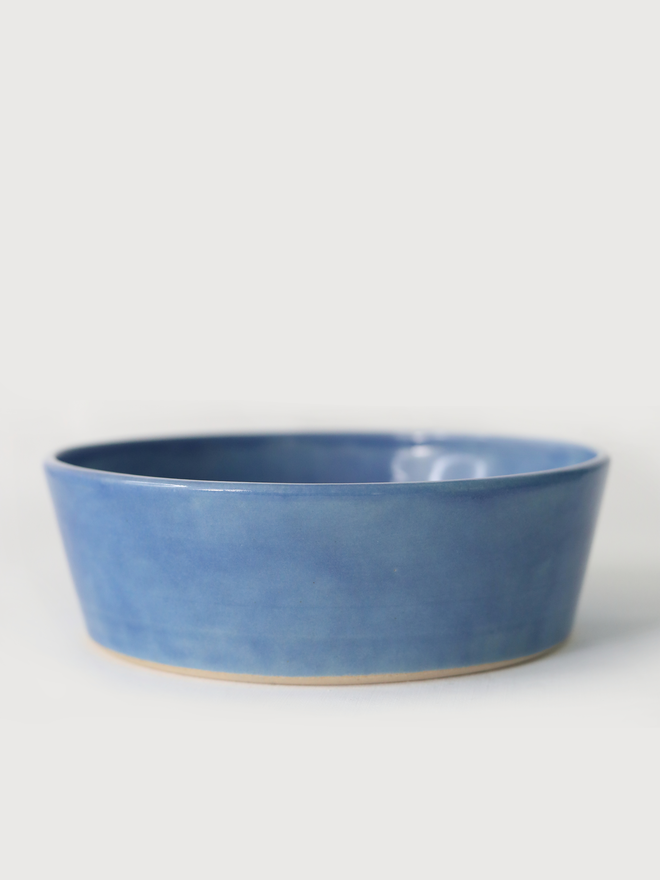 A single wide, shallow glazed ceramic pasta bowl in pacific blue, photographed from a slight angle against a plain light grey background, showing the unglazed natural clay base.