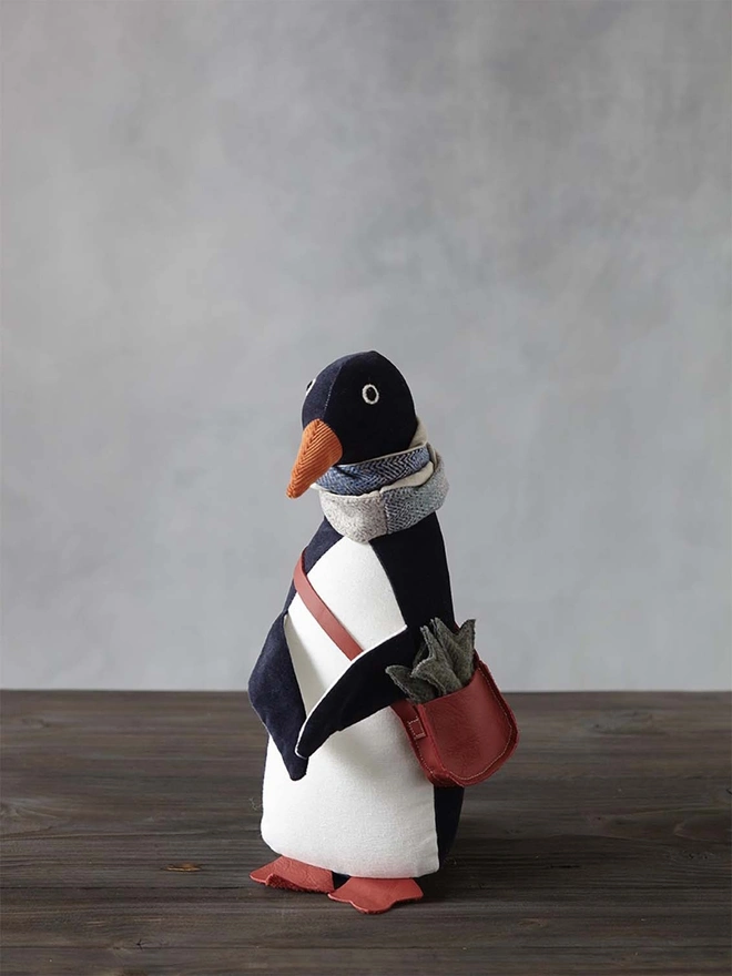 Dark blue velvet handmade toy penguin, wearing a tweed and linen scarf with a red leather satchel and three tweed fish