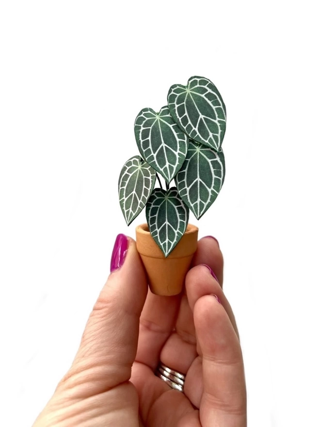 A miniature replica Anthurium Crystallinum paper plant ornament in a terracotta pot being held between 3 fingers against a white background