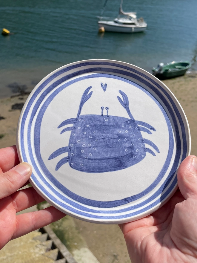 blue and white handmade stoneware plate with a blue handpainted happy crab with a little heart just between its claws. The plate has 3 blue lines around the rim.