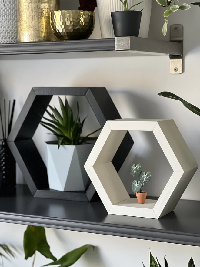 A miniature replica Philodendron Gloriosum paper plant ornament in a terracotta pot sat on a white hexagon shelf with a black hexagonal shelf to the left with a real plant inside, all sat on a large shelf with real plants and gold accessories in the background