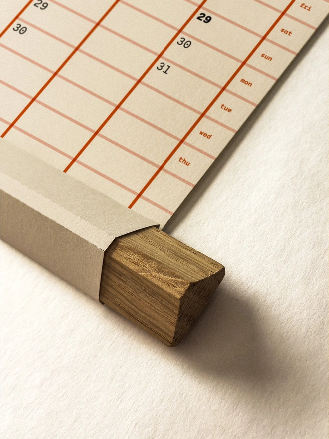 Closeup of yearly wall planner in orange on off-white paper, showing unique folded paper frame with oak strip insert.