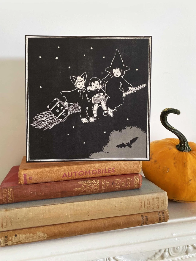 a halloween greeting card on a stack of books on a mantle piece featuring a vintage style illustration of a ghost girl and bat boy and vampire boy sat on a broom stick in the starry night sky