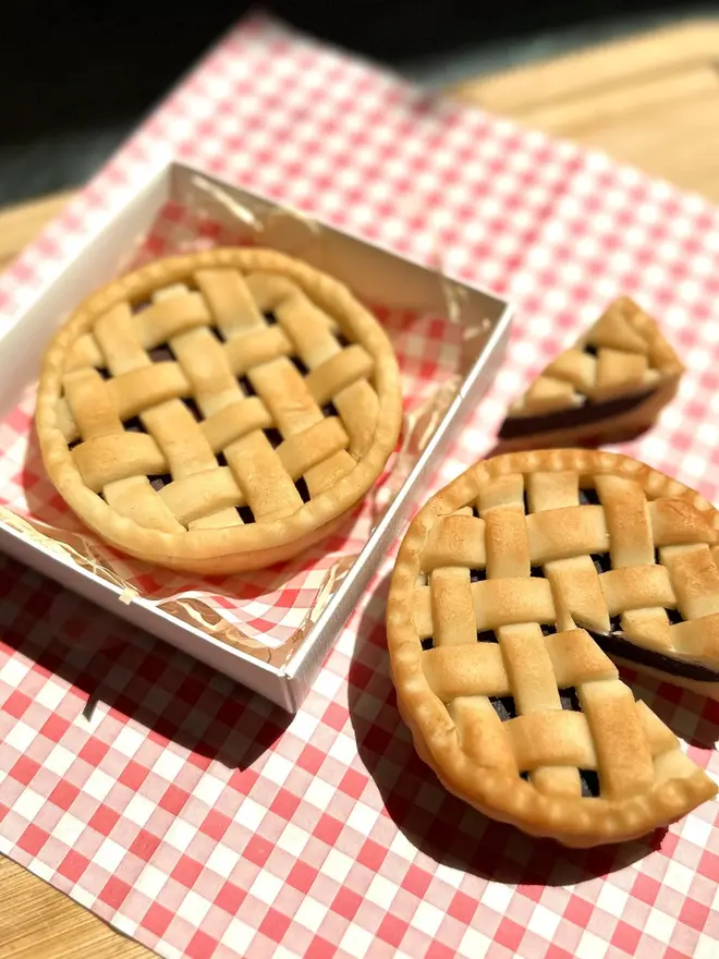 Marzipan Cherry Pie, a pair of marzipan cherry pies on a traditional picnic cloth. One is placed next to another that is packaged. 