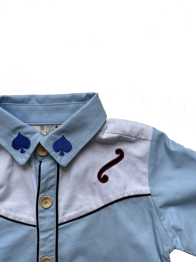 Detail of a blue and white cowboy shirt with navy piping and f-hole and spade detailing on the shoulder and collar.