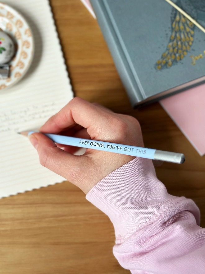 A hand holds a blue pencil with the words Keep Going, You've Got This along the side, and writes in a lined notebook on a wooden desk.