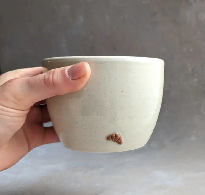 Vanilla bean–colored croissant coffee cup with a subtle croissant design, placed against a grey marble background.
