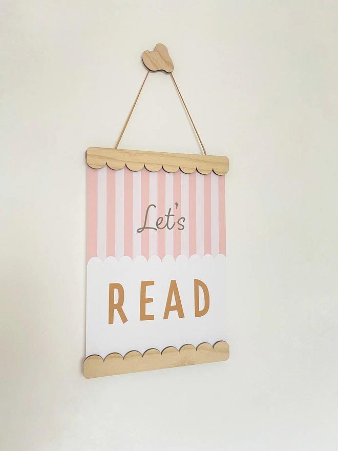A wooden magnetic picture frame, strung with vegan suede. Hung on a wavy wooden wall dot. Displaying a colourful stripy kids print that says Let’s Read 