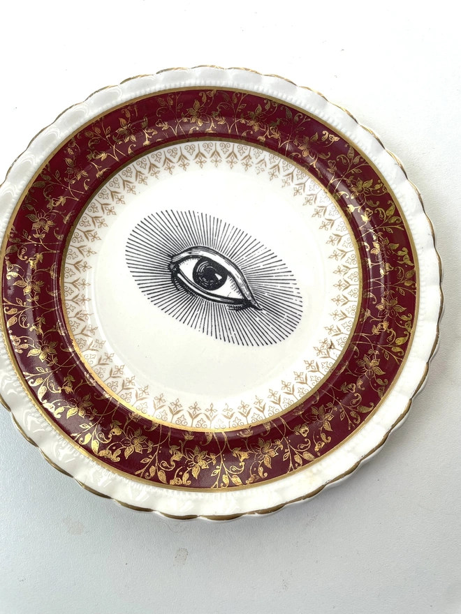 a really ornate red, gold and white china vintage plate has the vintage black and white image of an eye in the centre
