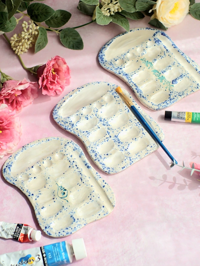 a trio of ceramic paint palettes, handcarved from stoneware clay and glazed in a splatter glaze with blue and white speckles. Each paint palette has a different design of bird, goddess or plain. Photographed with pink flowers on a pink background, surrounded by paints, Jenny Hopps Pottery, Ceramic gifts, artist gifts, painter gifts, creative gifts