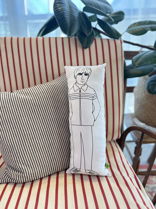 Black and white illustrated screen printed cushion of Oasis band member Noel Gallagher. The cushion sits on a striped sofa.