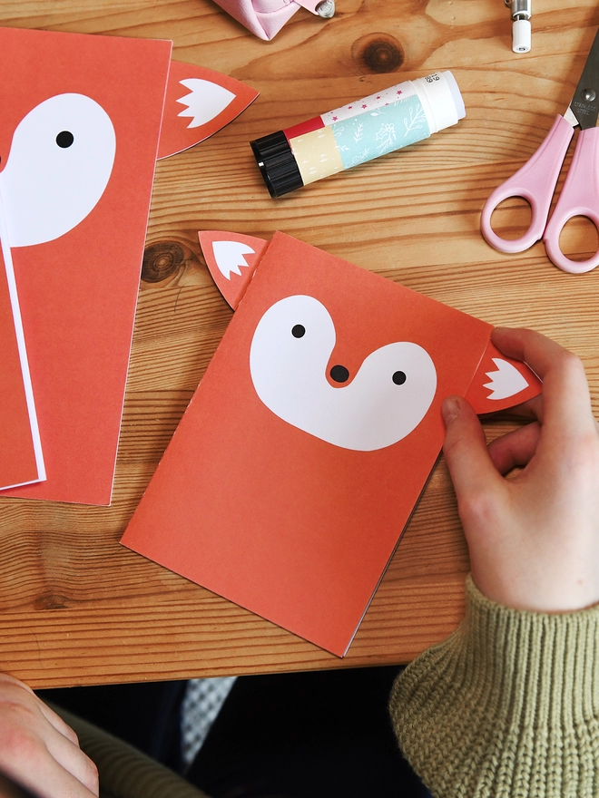 Several orange fox greetings cards lay on a wooden table. A child is gluing the ears to the corners of one card.