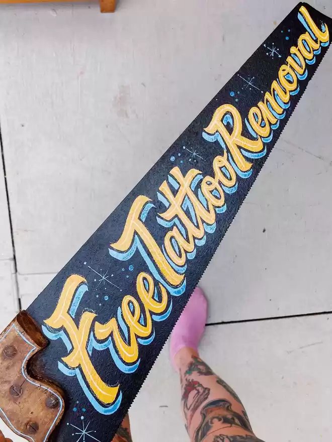 Hand-painted vintage saw spelling 'Free Tattoo Removal' in script with yellow and blues with sparkle details.