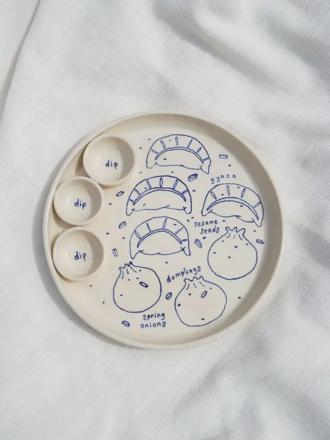 Dumplings Dippy Dinner Plate, a dipping plate sitting on a white bed cloth. It is covered in blue outlines for dumplings. 