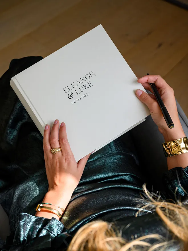 A pearl-coloured wedding guest book, personalised with printed lettering on the cover, rests on a woman's lap.