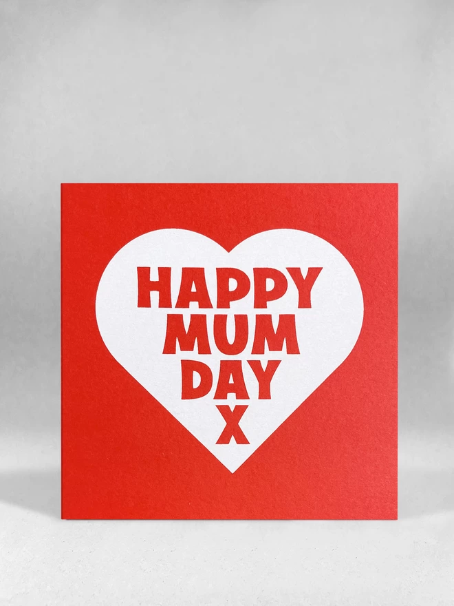 A red square card with a white heart, containing the words Mappy Mum Day x. Stood front on in a light grey studio 