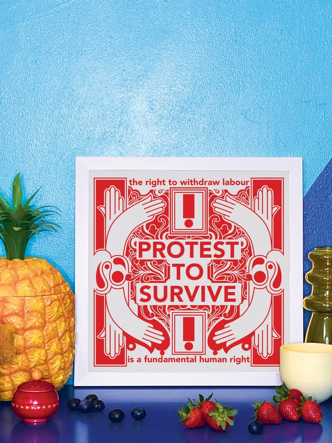 The bold red and white print with "Protest to survive" written at the centre, as well as “the right to withdraw labour” at the top, and “is a fundamental human right” at the bottom is in a white frame propped against a wall painted light and dark blue, on a shiny blue cabinet. Next to the frame is a plastic pineapple ice bucket, a small wooden red pot a handful of blueberries, a yellow glass vase, a small yellow ceramic pot and 4 strawberries.