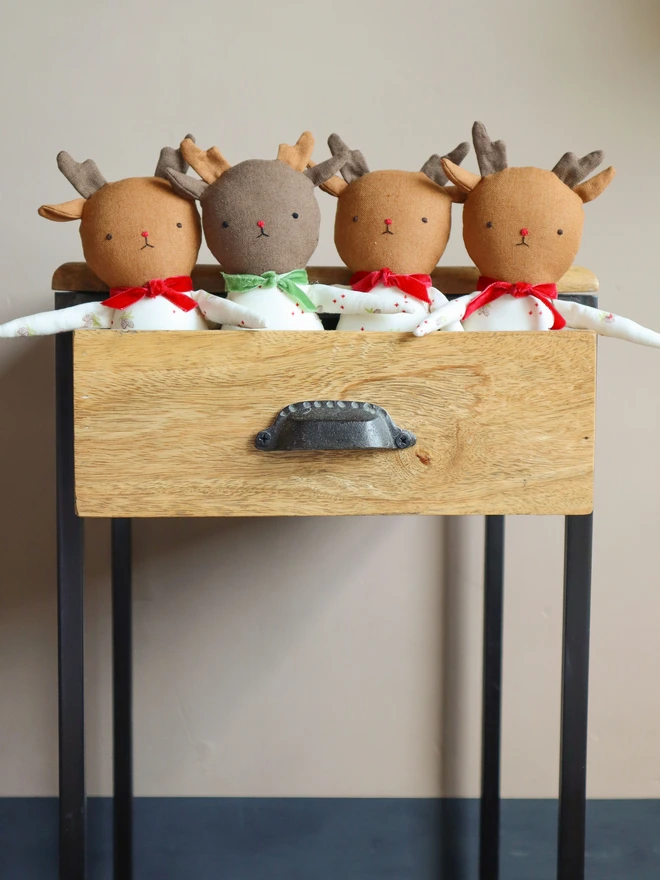 Four reindeer dolls arranged in a wooden drawer. Each doll has soft antlers, embroidered facial features, and a festive pinecone print outfit with either a red or green velvet bow. Two have green legs, and two have sparkly red legs. The dolls have "BOKUNO" fabric tags.