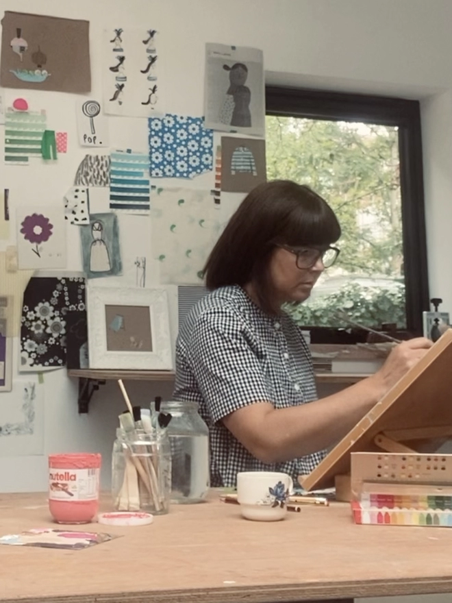 artist sitting at table strewn with art materials painting on drawing board