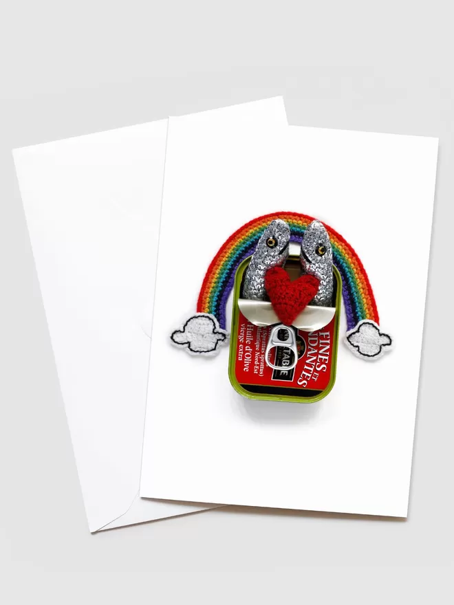Kate Jenkins Sardines Under a Rainbow Greetings Card. On the front of the card is a hand crotched rainbow with clouds at the bottom and sequinned sardines with a heart between them.