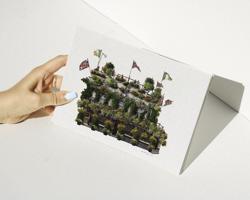 A hand with blue nail polish holding a print featuring an illustration of The Churchill Arms pub, heavily covered in flowers and flags, against a white background.