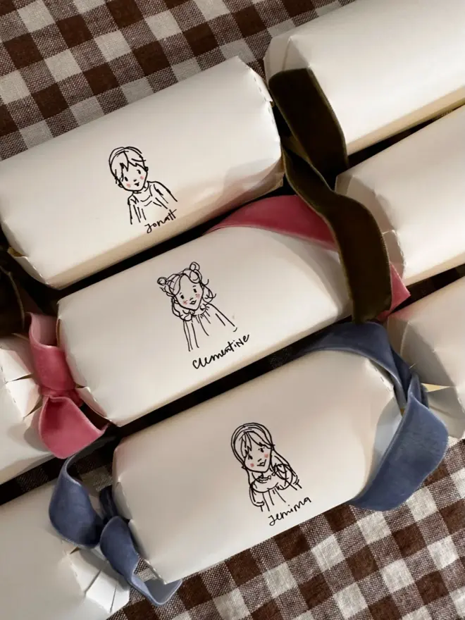 three white christmas crackers, each with a bespoke illustration of a child or pet, with different coloured velvet ribbon. sitting on brown and white gingham table cloth