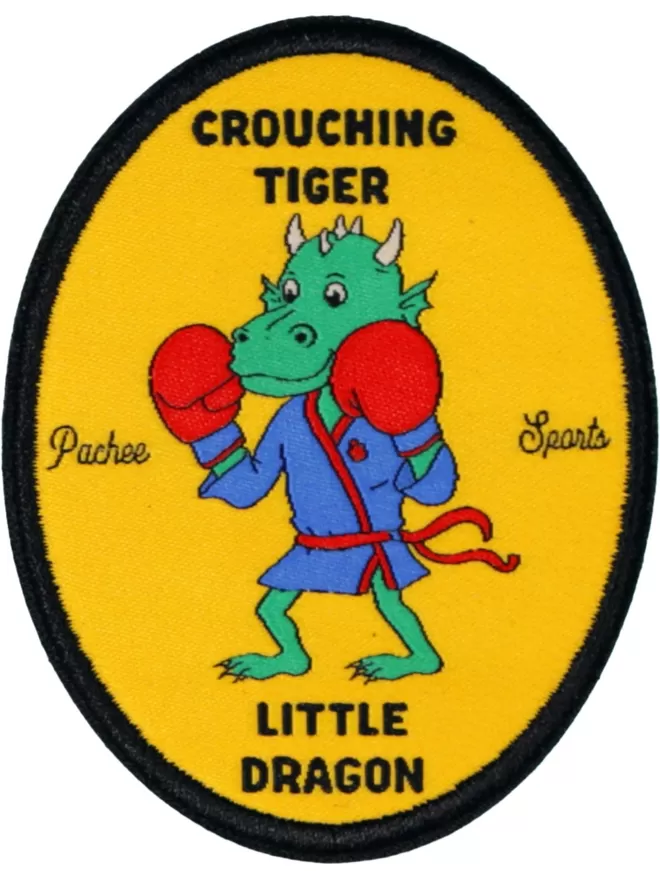 A yellow oval shaped patch with a horned dragon in the centre wearing boxing gloves. Crouching Tiger is written at the top and little dragon is written at the bottom.