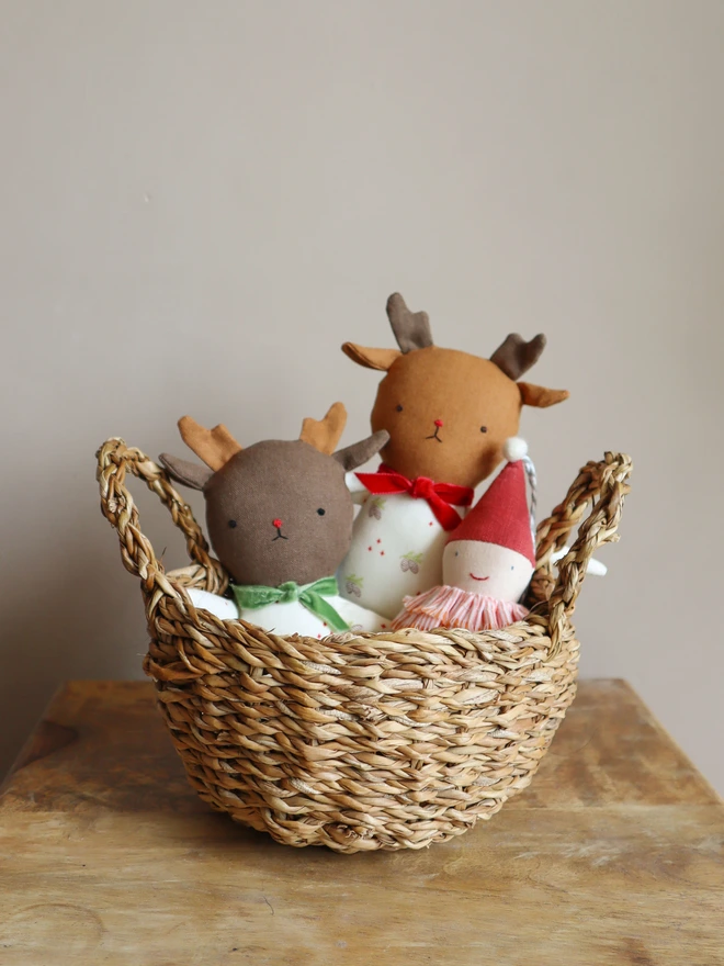 A woven basket on a wooden surface containing three handmade soft toys: two reindeer dolls with antlers and embroidered faces—one with a green bow, the other with a red bow—and a small smiling doll wearing a red pointed hat and striped collar. The basket is set against a plain beige background.
