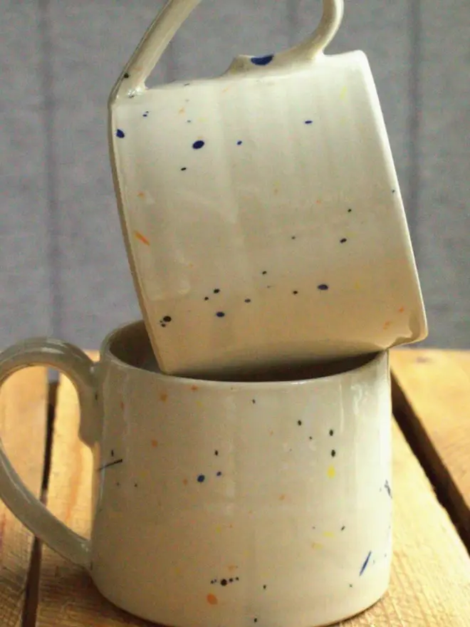 Two large creamy white terrazzo cups with black, blue, and yellow splatters and glossy glaze stacked on each other on a wooden surface.